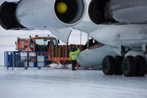 A worker in a high-visibility jacket unloads cargo from a large aircraft onto an icy surface in Antarctica, with an orange vehicle and stacked containers nearby—part of Antarctic Logistics and Expeditions operations.
