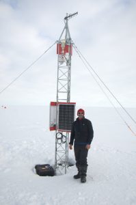 A person in winter clothing stands next to a tall, metal weather station with solar panels in snowy, flat Antarctica under a cloudy sky. The station, supported by guy wires, is part of Antarctic Logistics and Expeditions operations.