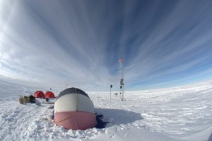 A snowy landscape in Antarctica with a round, dome-shaped tent in the foreground, two red tents nearby, boxes on the snow, and a tall weather station tower under a dramatic, streaked cloudy sky—perfect for Antarctic Logistics and Expeditions.