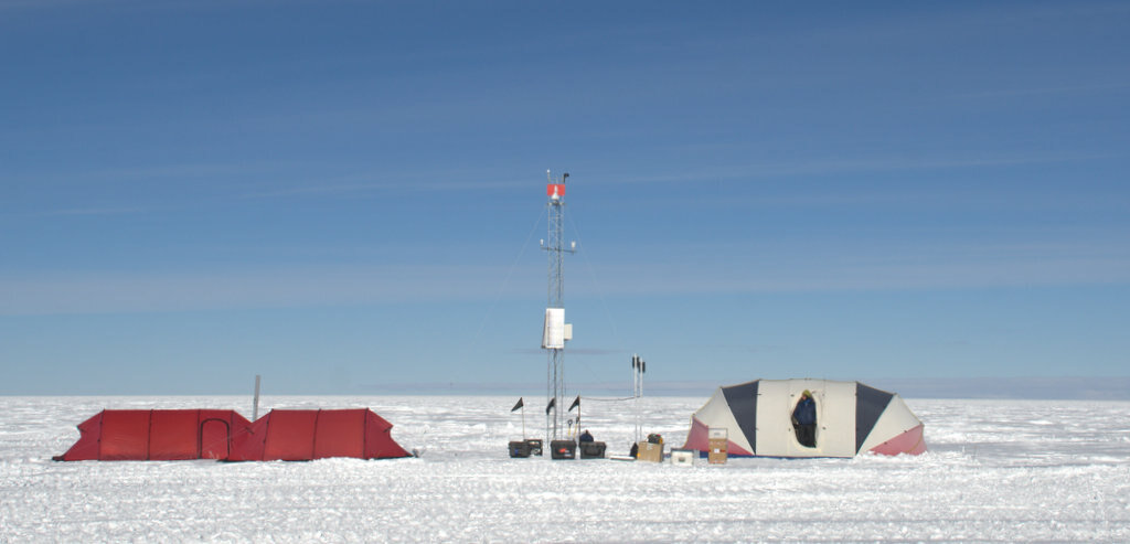 Two tents, one red and one gray, stand on a snowy landscape in Antarctica next to a tall weather station tower with instruments and flags. The sky is mostly clear with a few clouds near the horizon.