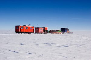 Bright red research station buildings and equipment are transported across the snowy expanse of Antarctica by a blue tracked vehicle under a clear blue sky, showcasing the vital work of Antarctic Logistics and Expeditions.