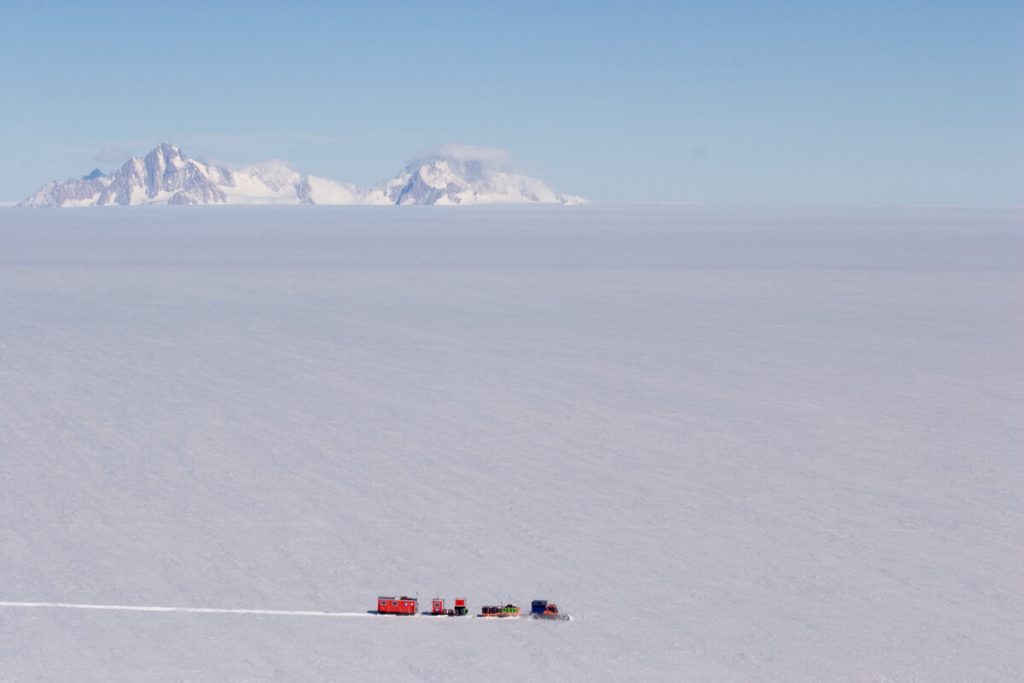 A small convoy of red vehicles from Antarctic Logistics and Expeditions travels across a vast, flat, snowy landscape with distant snow-covered mountains under a clear blue sky in Antarctica.