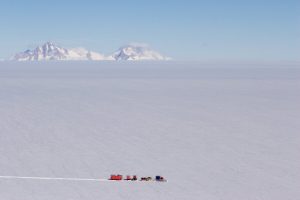 A small convoy of red vehicles from Antarctic Logistics and Expeditions travels across a vast, flat, snowy landscape with distant snow-covered mountains under a clear blue sky in Antarctica.