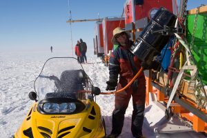 A person in winter clothing fuels a yellow snowmobile from a black barrel beside a mobile red structure on the snowy, flat landscape of Antarctica, with others working in the background under a clear blue sky.
