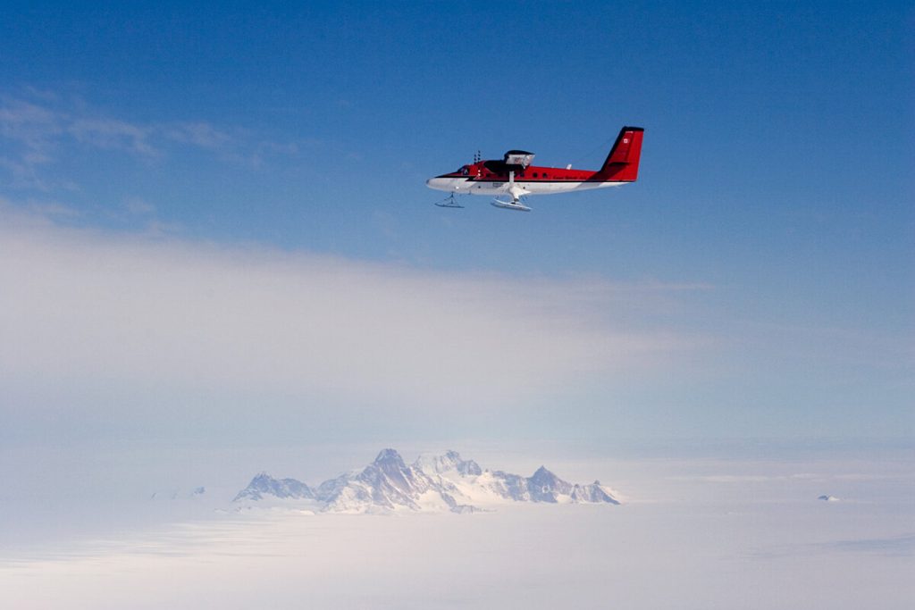 A red and white propeller airplane with skis, operated by Antarctic Logistics and Expeditions, flies over a vast snowy landscape in Antarctica, with a distant, rocky mountain range visible under a clear blue sky.