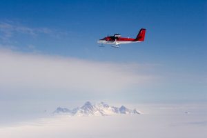 A red and white propeller airplane with skis, operated by Antarctic Logistics and Expeditions, flies over a vast snowy landscape in Antarctica, with a distant, rocky mountain range visible under a clear blue sky.