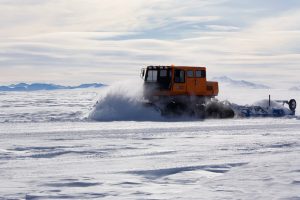 An orange snow vehicle from Antarctic Logistics and Expeditions drives through a snowy, frozen landscape near the South Pole, kicking up snow as it moves. Mountain peaks are visible in the distance under a cloudy sky.