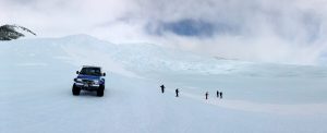 A blue off-road vehicle is parked on a vast snowy landscape in Antarctica, while five people walk across the snow under a cloudy sky, surrounded by white hills and icy terrain—part of an Antarctic Logistics and Expeditions journey.