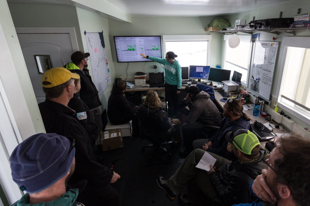 A group of people in a small office gather around as one person points to graphs on a large wall-mounted screen, analyzing data from Antarctic Logistics and Expeditions, with computers, notes, and equipment visible around the room.