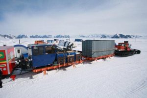 A tracked vehicle with sledges loaded with containers and supplies traverses the snowy, icy landscape of Antarctica, mountains rising in the distance beneath a cloudy sky—a scene typical for Antarctic Logistics and Expeditions.
