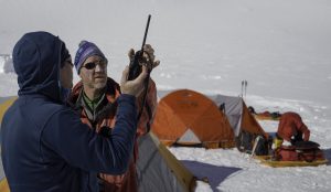 Two people in winter gear use a walkie-talkie at a snowy campsite near the South Pole, with orange tents and equipment in the background. The area, managed by Antarctic Logistics and Expeditions, is blanketed in snow under bright sunlight.
