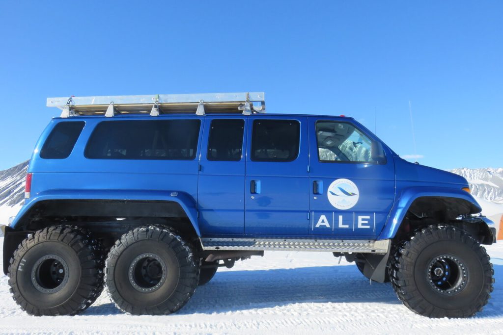 A large blue six-wheeled off-road vehicle with the ALE (Antarctic Logistics and Expeditions) logo parked on snow, with mountains and a clear blue sky in the background, ready for an adventure in Antarctica.