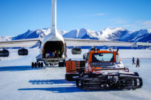 A cargo plane with its rear open is being unloaded on an icy runway in Antarctica, with snowy mountains in the background and an orange Antarctic Logistics and Expeditions snow vehicle parked nearby.
