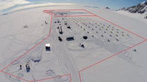 Aerial view of a snowy Antarctic Logistics and Expeditions research camp with rows of tents and buildings outlined by a red border. Snow-covered landscape and mountains are visible in the background under a clear South Pole sky.