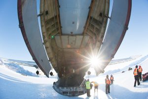 A large cargo plane with its rear ramp open sits on snowy ground in Antarctica. Several workers in orange and yellow safety vests, supporting Antarctic Logistics and Expeditions, stand nearby under a bright sun and snow-covered mountains.
