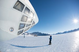 A person stands on a vast, snowy landscape near a large, white aircraft nose in Antarctica. Distant mountains and the bright sun shine under a clear blue sky, with another person and Antarctic Logistics and Expeditions vehicles in the background.