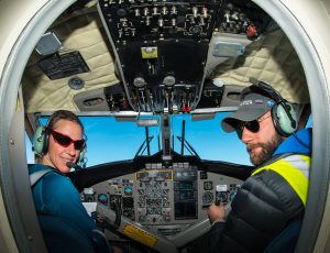 Two pilots wearing headsets and sunglasses sit in the cockpit of an Antarctic Logistics and Expeditions airplane, surrounded by control panels, looking back toward the camera with blue sky visible through the windows en route to the South Pole.