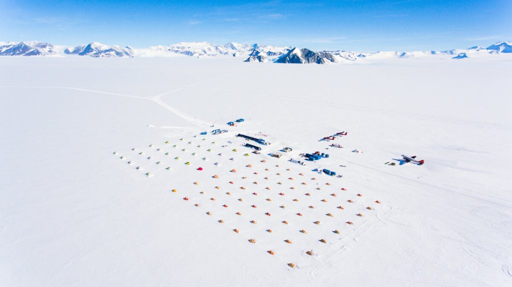 Aerial view of an Antarctic Logistics and Expeditions research camp on a vast snowfield, with colorful tents in neat rows, small buildings, and two aircraft. Snow-covered mountains rise in the distant background under a clear blue sky.