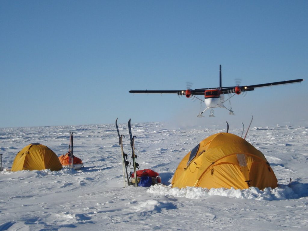 A small airplane flies low over a snowy Antarctic landscape with three yellow-orange tents and ski equipment set up in the foreground under a clear blue sky, ready for an Antarctic Logistics and Expeditions adventure.