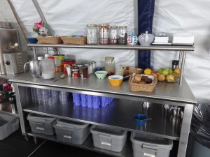A stainless steel kitchen counter with jars, baskets, canned goods, fruit, bowls, cups, utensils, and storage bins underneath sits in a tented area—perfect for Antarctic Logistics and Expeditions at the South Pole.