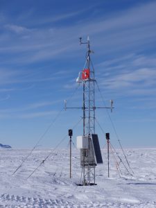 A weather station with sensors, solar panels, and cables stands on a snowy, flat landscape in Antarctica under a blue sky with wispy clouds. Mountains are visible in the distant background, highlighting Antarctic Logistics and Expeditions.