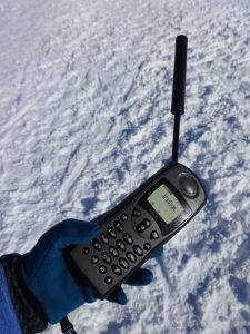 A gloved hand holds an old-fashioned satellite phone with an antenna, displaying “Iridium” on its screen, against a snowy South Pole backdrop, highlighting vital communications in Antarctic Logistics and Expeditions.