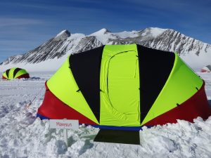 A neon green and black tent stands in snowy terrain with mountains in the background, marking an expedition in Antarctica. A sign labeled Mawson rests in the snow, while other tents from Antarctic Logistics and Expeditions are visible in the distance.
