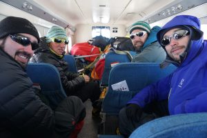 Four men wearing winter gear and sunglasses sit inside a small plane with blue seats and gear piled in the back, smiling at the camera en route to the South Pole with Antarctic Logistics and Expeditions.