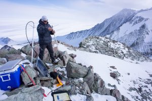 A person in winter gear sets up scientific equipment atop a rocky, snow-covered mountain ridge in Antarctica, with solar panels and wires, surrounded by snowy peaks under a cloudy sky—an expedition reminiscent of Antarctic Logistics and Expeditions.