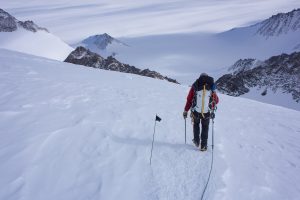 A climber in winter gear and a backpack walks up a snowy mountain slope in Antarctica, following a rope past a small black flag, with rugged peaks and the vast Antarctic landscape stretching behind him.