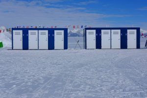 A row of blue and white portable toilets stands on snowy ground in Antarctica, with mountains in the background and flags from various countries strung above—part of Antarctic Logistics and Expeditions operations near the South Pole under a partly cloudy sky.