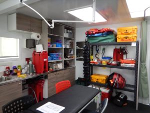 A well-organized medical room at the South Pole with cabinets, shelves holding emergency supplies, equipment, and first aid kits, a padded exam table, chairs, and a countertop with a sink. Bright lighting and a clean, orderly appearance.