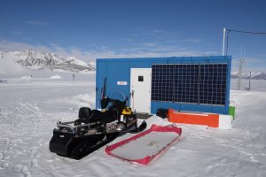 A blue research station with solar panels stands in Antarctica’s snowy landscape. In front, a snowmobile, red sled, and equipment await. Snow-covered mountains and a clear blue sky complete this South Pole scene.
