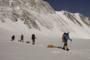 Four climbers in winter gear are pulling sleds with supplies across a snow-covered Antarctic landscape, with steep mountain slopes in the background under a clear blue sky—a classic scene of South Pole expeditions.
