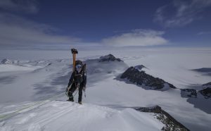 A mountaineer in cold-weather gear stands on a snowy peak in Antarctica, skis strapped to their backpack and attached to a safety rope, with vast Antarctic Logistics and Expeditions vistas and a cloudy sky in the background.