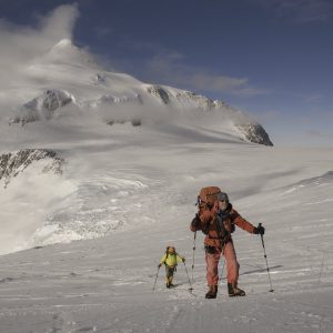 Two climbers trek up a snowy, icy mountain slope in Antarctica under a blue sky, wearing bright winter gear and carrying backpacks, with a snow-covered peak—reminiscent of Antarctic Logistics and Expeditions—in the background.