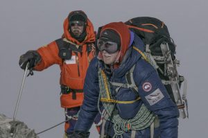 Two climbers in heavy winter gear, goggles, and backpacks ascend a snowy, foggy mountain in Antarctica. One is in a red jacket holding a pole; the other in blue with rope coiled around their chest. Visibility is low—true South Pole conditions.