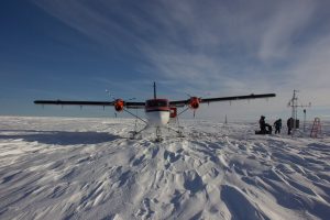 A small red and white airplane with skis, operated by Antarctic Logistics and Expeditions, lands on a snowy flat landscape in Antarctica. Several people work near scientific equipment beside the plane under a clear blue sky.