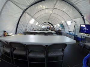A large, white tent at the South Pole features rows of folding tables and chairs set up for a meeting. A whiteboard, a monitor displaying an airplane, and supplies on a side table reflect Antarctic Logistics and Expeditions in action. People stand in the distance.