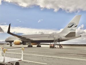 A white passenger airplane with the “Antarctic Logistics and Expeditions” logo is parked on the tarmac. Several people in orange vests stand near portable stairs by the aircraft under a partly cloudy sky, preparing for an Antarctica journey.