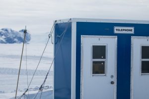 A blue and white building labeled Telephone stands on a snowy, icy landscape near the South Pole, with mountains in the background under a cloudy sky—part of Antarctic Logistics and Expeditions.
