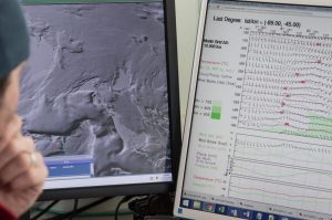 A person looks at two computer monitors displaying satellite imagery of Antarctic terrain on the left and detailed weather data charts with graphs and wind readings from the South Pole on the right.
