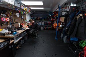 A person sits at a workbench in a well-organized storage and workspace filled with shelves, gear, tools, bins, and outdoor equipment—perfect for prepping Antarctic Logistics and Expeditions to the South Pole. The area is brightly lit with overhead lights.