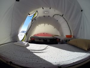 Inside view of a white tent in Antarctica with two beds, a folded towel on one, a pillow on the other, and a small table with an orange bowl. Daylight streams in from the open entrance as snow blankets the landscape outside—true South Pole style.
