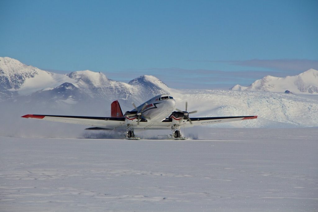 A propeller plane with skis lands on a snowy, flat expanse in Antarctica, surrounded by snow-covered mountains under a clear blue sky—another day for Antarctic Logistics and Expeditions near the South Pole.