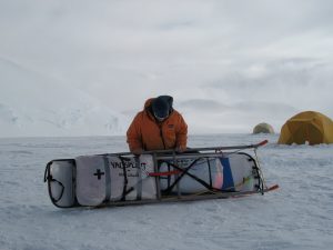 A person in an orange parka and winter gear stands in snowy conditions, securing gear onto a sled. Two yellow tents are set up in the background with snow-covered mountains—an Antarctic Logistics and Expeditions journey.
