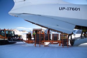 Workers unload cargo from the open rear of a large aircraft onto sleds, with snow vehicles nearby on an Antarctic landscape surrounded by mountains under a clear sky.