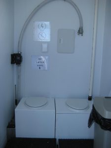 Two white toilet seats with closed lids sit side by side in a South Pole restroom. A sign above reads NO PAPER HERE. A hose runs above the toilets, and there is a trash can with a black liner on the right.
