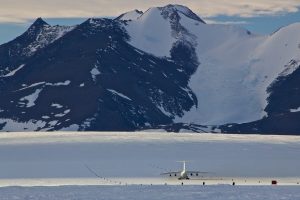 A large aircraft sits on a snowy runway surrounded by snow-covered mountains and rugged dark peaks under a partly cloudy sky in the remote, icy landscape of Antarctica.