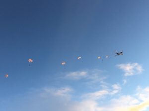 A plane flies through a blue sky over Antarctica while several parachutists, each with an open orange parachute, descend in a line behind it. Fluffy clouds are scattered below—part of an Antarctic Logistics and Expeditions adventure.